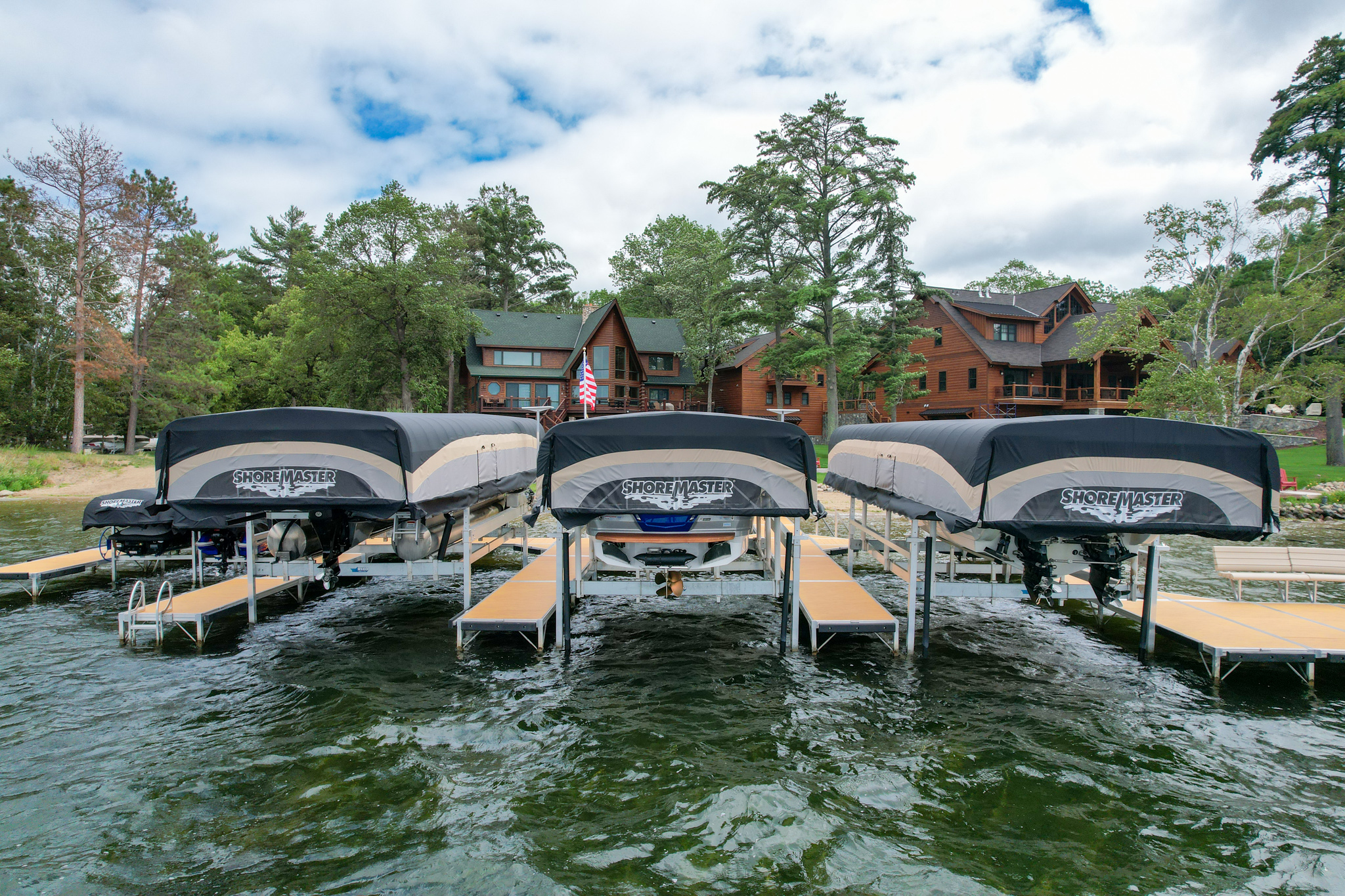 Three covered ShoreMaster boat lifts and a dock system sit in the water in front of two large lakeside houses surrounded by lush green trees.