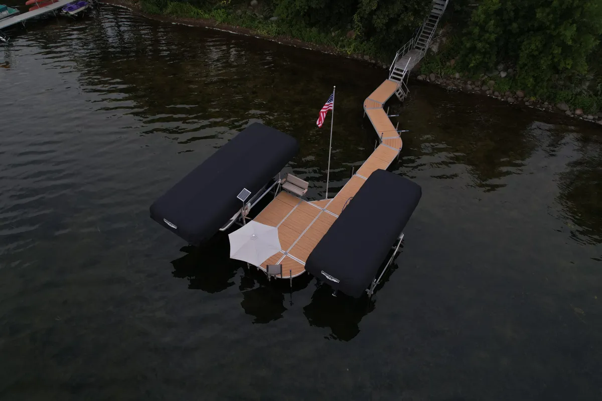 Aerial view of a custom lakeside dock with two covered boat lifts, wooden decking, an American flag, and shoreline stairs.