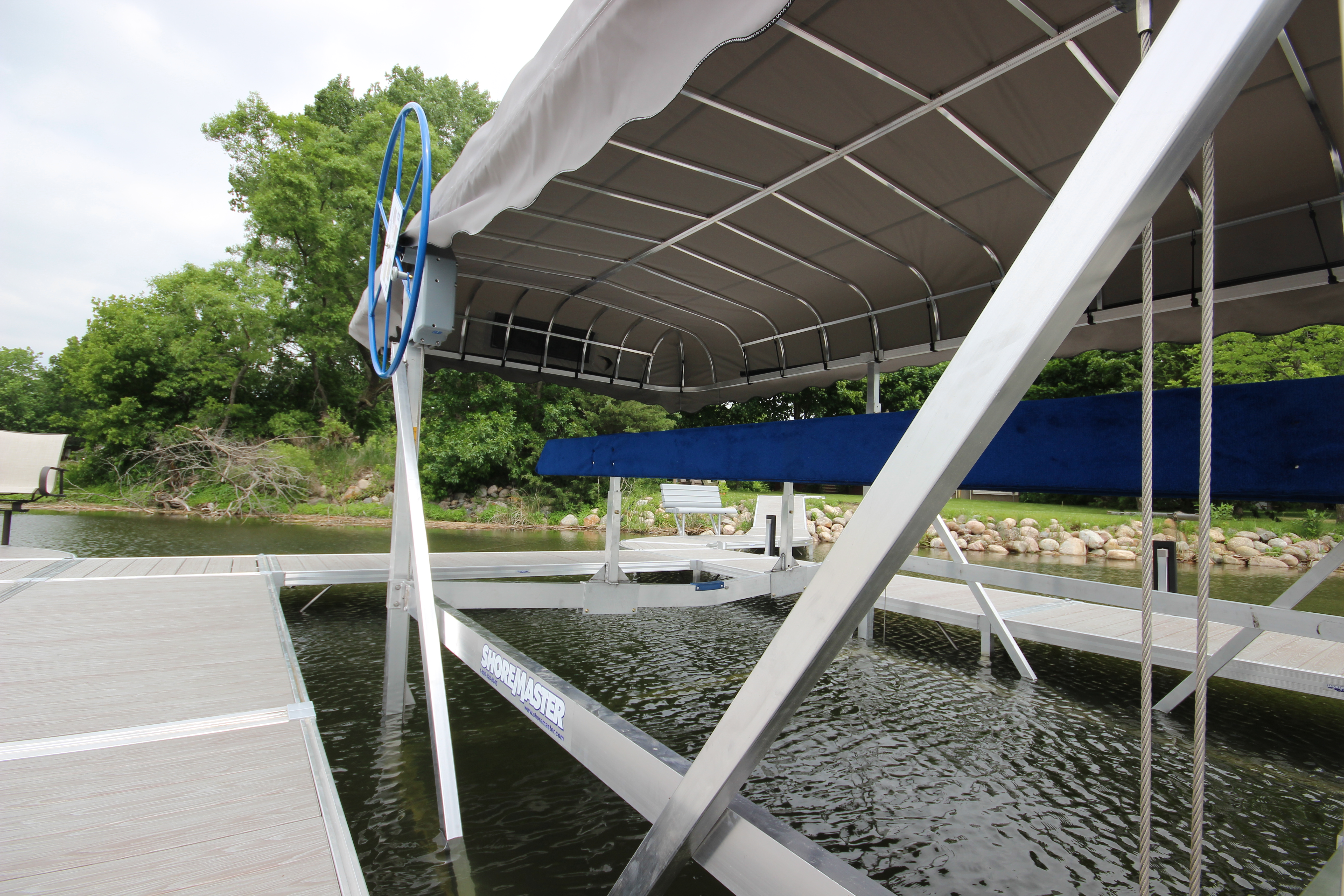 A ShoreMaster boat lift with a canopy is installed on a dock next to a body of water, featuring a blue hand wheel and blue bunks, with trees in the background.