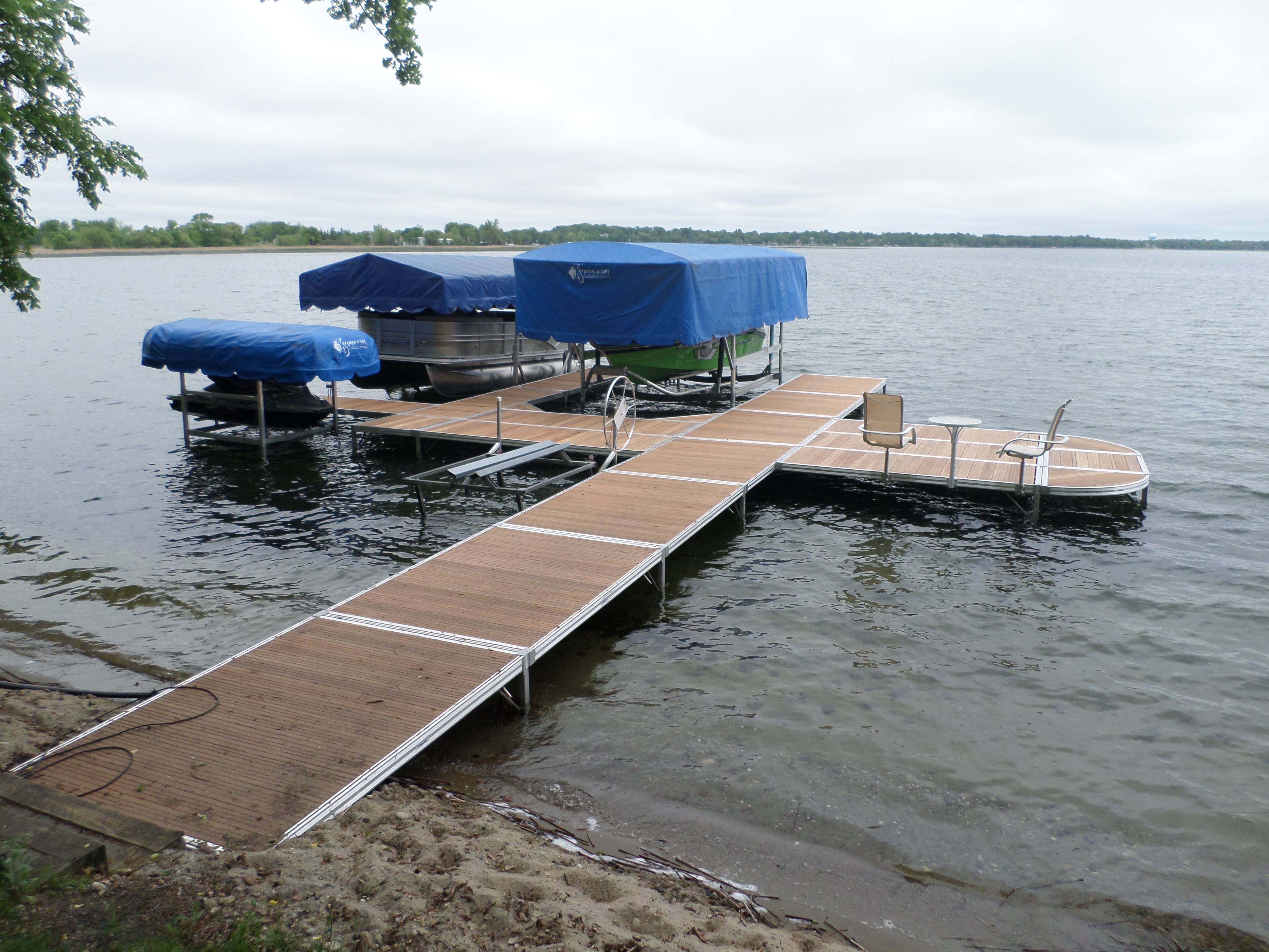 A dock with two boat lifts and blue canopies extending into a lake. There are two boats on the lifts, and some furniture on the dock extension.