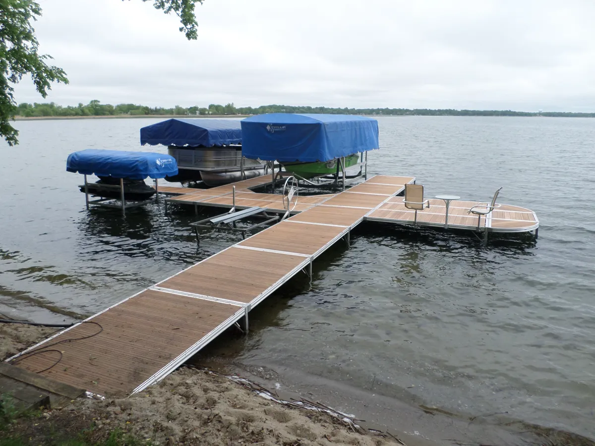 A dock with two boat lifts and blue canopies extending into a lake. There are two boats on the lifts, and some furniture on the dock extension.