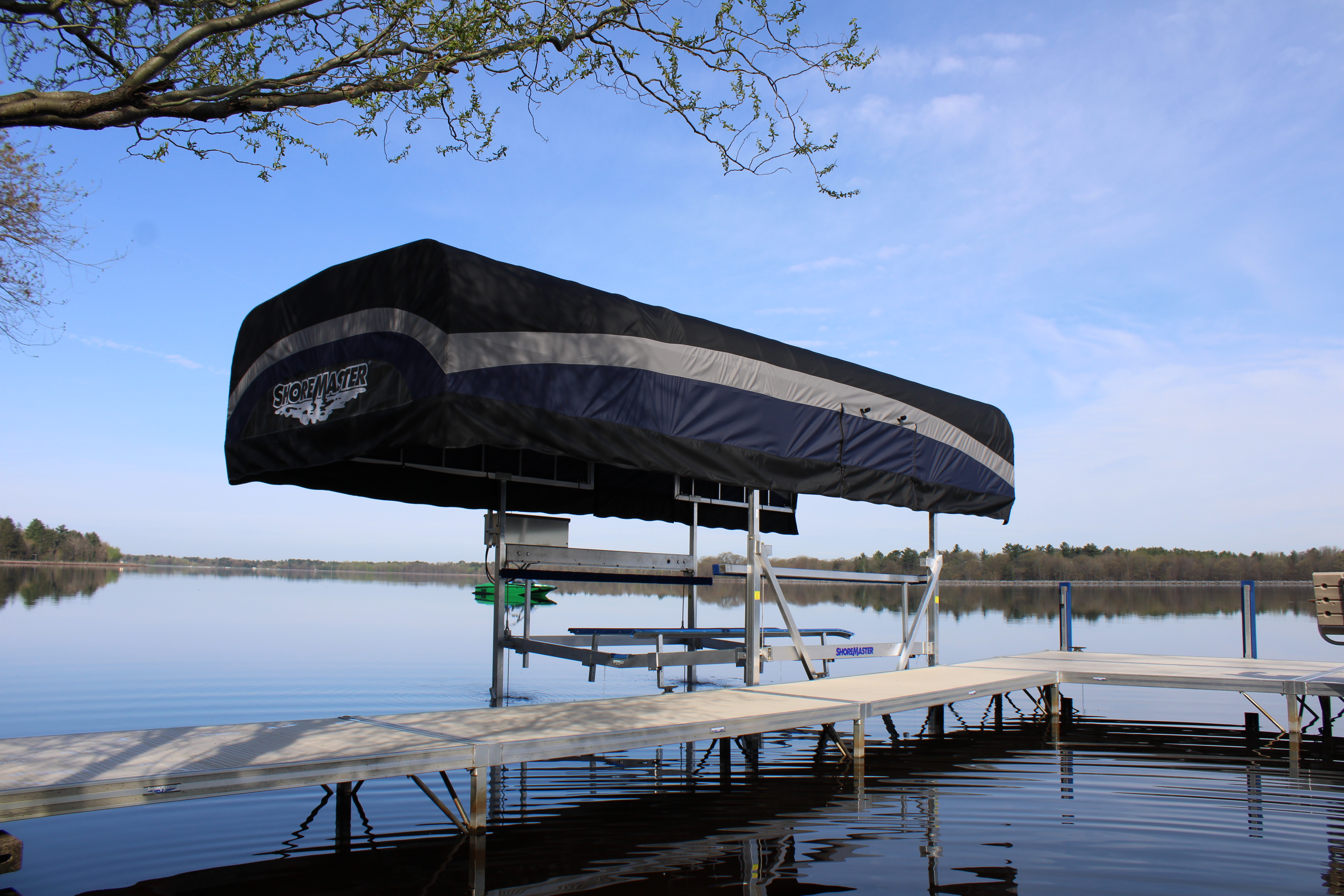 ShoreMaster covered boat lift beside an aluminum dock on a calm lake under a clear blue sky.